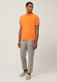 Narančasta polo majica, svijetlosivi hlače s uzorkom i svijetlosivi mokasini. Hlače imaju botanički uzorak. Jednostavan, ležeran outfit.