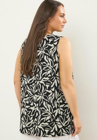 Mujer con cabello largo y marrón, vestida con una blusa negra sin mangas con patrones de hojas blancas, de pie de perfil contra un fondo claro y liso.