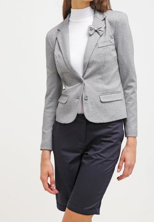 Woman wearing a gray fitted blazer with bow detail, white turtleneck, and dark knee-length shorts, standing against a plain background.
