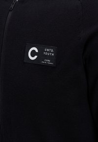 Svart zip-up tröja med texturerad tyg, logotygmärke med vit text och ribbad detalj längs kanterna.