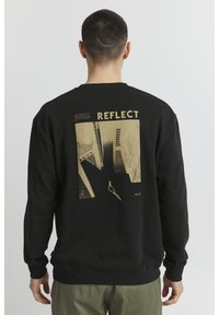 Čierna mikina s veľkým zlatým grafickým potlačou na chrbte so slovom "REFLECT" a abstraktnými dizajnmi budov.