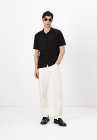 Camisa negra de manga corta con textura de patrón de diamante, cierre frontal de botones, combinada con pantalones blancos y zapatos negros. Diseño minimalista.