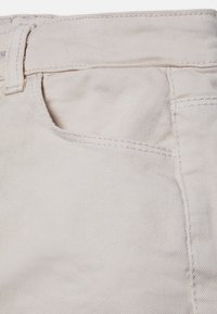 Tecido de denim bege, com um bolso frontal, costura visível e uma superfície texturada e ribada. Vista em close, demonstrando os detalhes do design.