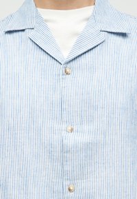 Κοντινό πλάνο μπλε και λευκού πουκαμίσου με κάθετες ρίγες και κουμπιά που μοιάζουν με πέρλες πάνω από λευκό εσωτερικό πουκάμισο, δείχνοντας γιακά και περιοχή στήθους.