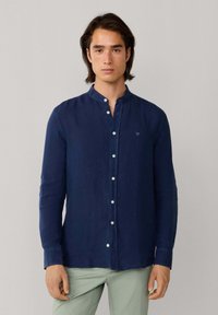 Homme portant une chemise à boutons bleu marine avec un col mao et un pantalon vert clair, debout devant un fond gris uni.