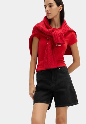 Kvinde iført en rød t-shirt med en rød kabelstrikket sweater draperet over skuldrene og sorte denimshorts, kigger til siden.