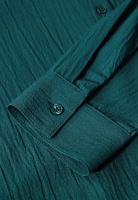Camicia teal con tessuto texturizzato e motivo a righe verticali, polsino con bottoni e dettaglio di bottone coordinato.