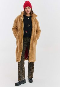 Žena v dlouhém béžovém teddy kabátu, černém svetr s výšivkou houbou, kalhoty s leopardím potiskem, černých kozačkách a červené pletené čepici, stojící.