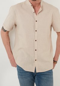 Beige overhemd met korte mouwen en een mandarinakraag, voorzien van bruine knopen. Gemaakt van textuurstof, heeft een rechte snit en een subtiel geweven patroon.