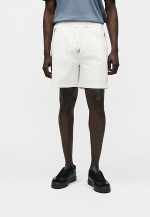 Man met witte shorts met trekkoord, zwarte sneakers en een blauw overhemd, staand met ontspannen armen tegen een witte achtergrond.