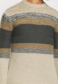 Homme portant un pull en tricot avec des rayures horizontales beige, marron, gris foncé et crème.