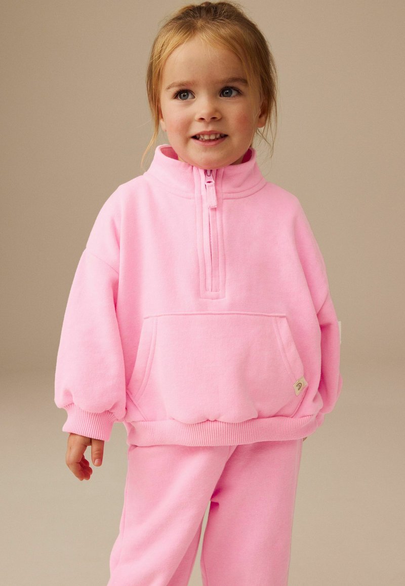Joven niña sonriendo, vestida con una sudadera rosa con cremallera y pantalones a juego, sobre un fondo beige liso.