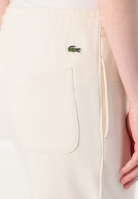 Krämfärgade mjukisbyxor i en strukturerad textil, med en sidficka och en liten, grön broderad krokodil-logga på baksidan.