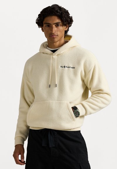 Sudadera de felpa crema con bolsillo canguro, capucha ajustable con cordón y logo bordado en negro. Combinada con pantalones negros y un reloj.