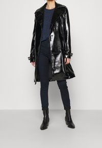 Zwarte glanzende trenchcoat met brede revers, knoopsluiting en gespaccents, gecombineerd met een blauw patroon top en donkere jeans.