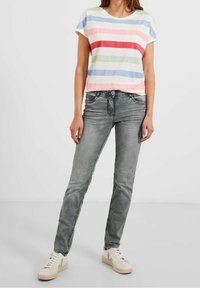 Woman wearing a short-sleeve striped pastel shirt, gray faded jeans, and white sneakers standing against a plain light background.