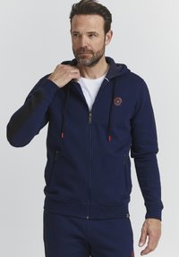 Marineblaue Zip-Hoodie aus strukturiertem Stoff, mit verstellbaren Kordeln, orangefarbenen Akzenten und einem Logo auf der Brust. Seitentaschen sind ebenfalls vorhanden.
