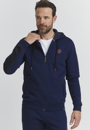 Navy blue zip-up hoodie with a textured fabric, adjustable drawstrings, orange accents, and a logo on the chest. Side pockets included.