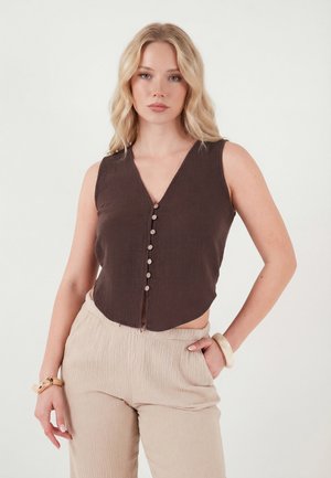 Szőke nő, ujjatlan sötétbarna gombos felsővel és bézs texturált nadrággal, mindkét csuklóján karkötővel, egyik kezét zsebében tartva áll.