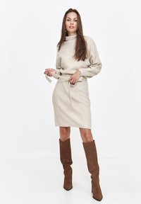 Beige gestrickter Pullover mit hohem Kragen und einem passenden Bleistiftrock, der an der Taille mit einem Gürtel tailliert ist. Braune kniehohe Stiefel aus Wildleder.