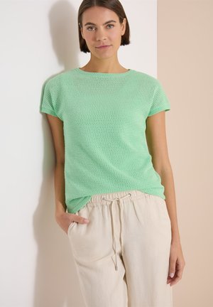 Femme aux cheveux bruns courts debout contre un mur, portant un haut en maille vert clair et un pantalon beige à cordon, une main dans la poche.