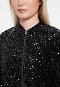 Czarna bomberka pokryta cekinami, z zapięciem na zamek błyskawiczny, wysokim kołnierzem oraz teksturowaną powierzchnią, podkreślająca blask i błysk.