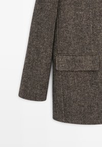 Brown tweed blazer with a textured finish, single pocket on the left-front, and tailored silhouette. Visible stitching along the seams.