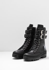 Botines negros de cuero con cordones en la parte delantera, herrajes metálicos, cremallera lateral, dos correas y suela de goma texturizada con relieves.