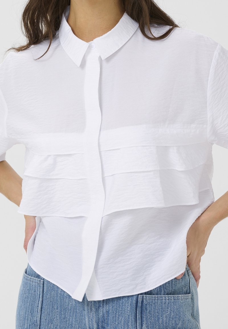 Kvinde iført en hvid kortærmet skjorte med lagdelte flæser og krave, parret med høj talje blå denim jeans.