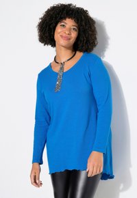 Blaues Langarmoberteil mit rundem Ausschnitt, strukturiertem Stoff und asymmetrischem Saum. Kombiniert mit glänzenden schwarzen Leggings für einen Kontrast.