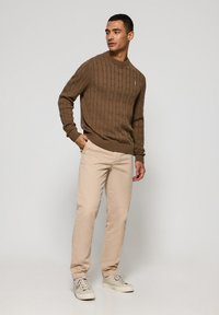 Suéter marrón de punto trenzado con cuello de canalé, combinado con pantalones beige claros y zapatillas beige. El modelo está de pie frente a un fondo neutral.