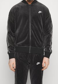 Svart sammets zip-up jacka med vita ränder. Har en ribbad krage och muddar samt en liten vit Nike-logotyp på bröstet.