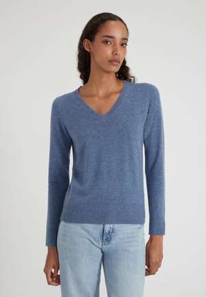 Junge Frau in einem blauen V-Ausschnitt-Pullover und hellblauen Jeans, die vor einem schlichten weißen Hintergrund steht.