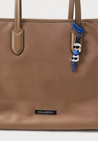 Καφέ τσάντα χειρός με δύο λουριά ώμου, κατασκευασμένη από λείο υλικό. Διαθέτει διακοσμητικό γούρι με μπλε και μαύρο σχέδιο και πλακέτα με το λογότυπο.