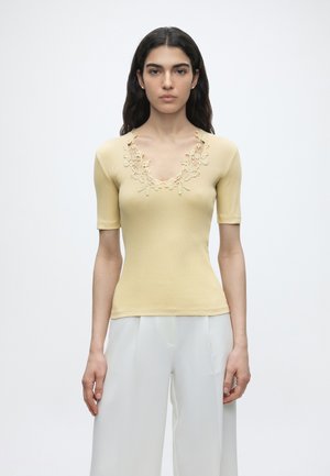 Frau trägt ein hellgelbes kurzärmeliges Oberteil mit Spitzen-V-Ausschnitt und weiße Hosen mit weitem Bein, steht vor einem schlichten weißen Hintergrund.