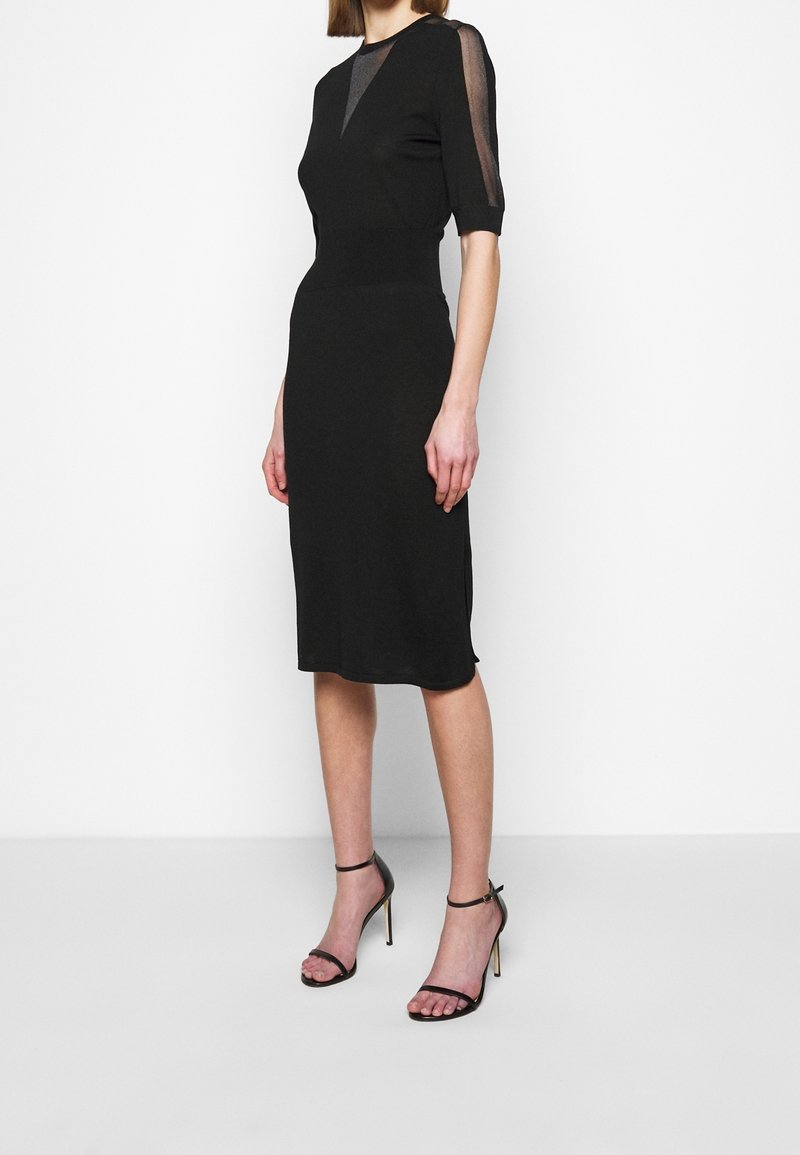 Femme portant une robe noire longueur genou avec des manches courtes transparentes et des sandales noires à talons hauts avec des brides à la cheville, debout sur un sol blanc.