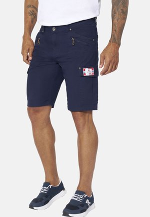 Mann trägt marineblaue Cargoshorts mit Reißverschlüssen und Klappentaschen, weißes T-Shirt, blau-weiße Sneakers und sichtbare Armtattoos.