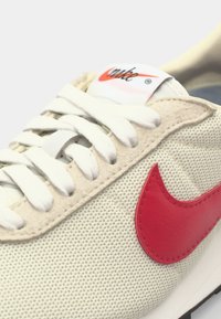 Nike sneakers with a perforated light beige fabric upper, white laces, and a prominent red leather swoosh logo. Textured accents noted.