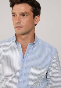 Camisa de botones a rayas azul y blanca con un bolsillo en el pecho y un cuello suave, hecha de tela tejida, que muestra delgadas rayas verticales.