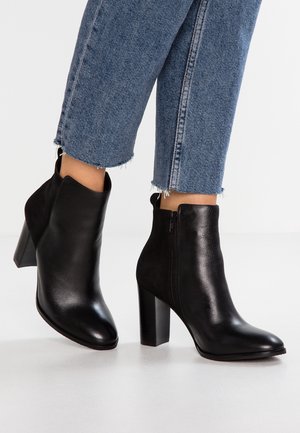 Botines de cuero negro con punta afilada y tacón alto y chunky. Cierre con cremallera lateral. Llevados con jeans azules de dobladillo deshilachado.