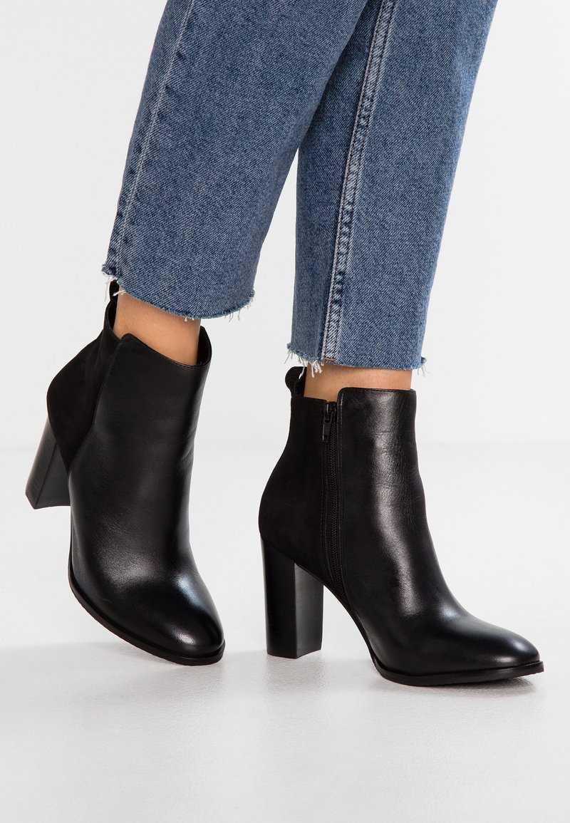 Botines de cuero negro con punta afilada y tacón alto y chunky. Cierre con cremallera lateral. Llevados con jeans azules de dobladillo deshilachado.