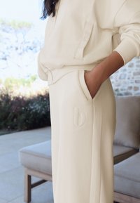 Cream-colored sweatshirt and matching pants in soft fabric. Pants feature side pockets and an embroidered 'b' on the hip. Relaxed fit.