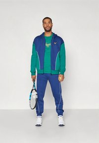 Homem em fato de treino azul e verde segurando uma raquete de ténis e uma bola, de pé contra um fundo claro.