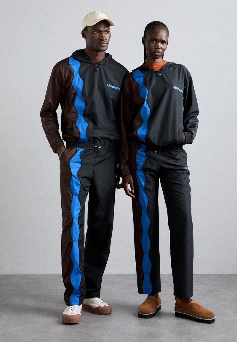 Survêtements noirs et marron avec des motifs ondulés bleus, présentant des capuches et des ceintures à cordon de serrage. Les modèles portent des chaussures décontractées et une casquette.
