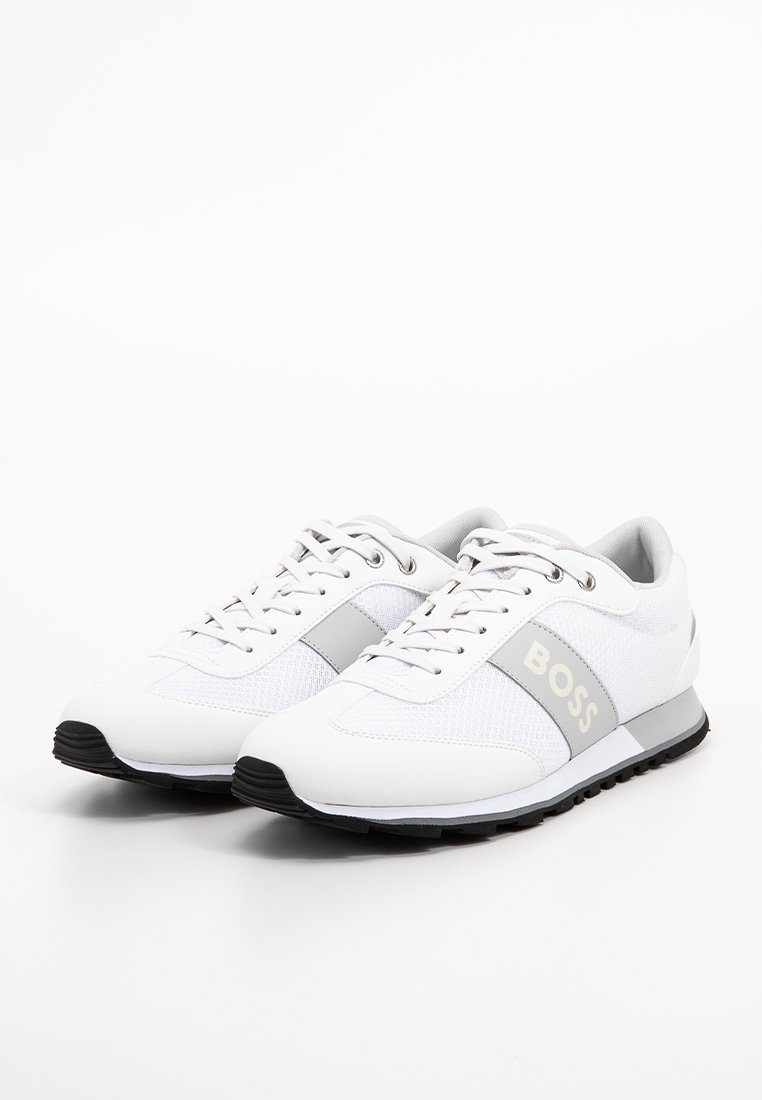 Un par de zapatillas blancas de corte bajo con el logo gris "BOSS", cordones blancos y suelas texturizadas negras, mostradas sobre un fondo blanco.