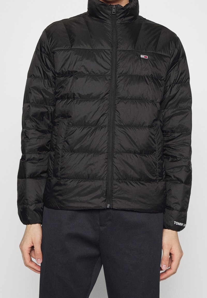 Homme portant une veste noire matelassée zippée avec un petit logo sur la poitrine et un texte de marque sur le poignet, associée à un pantalon foncé.
