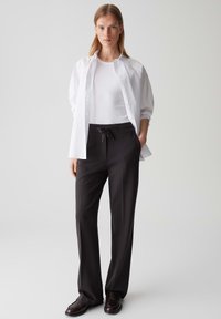 Camicia bianca a pulsante sopra una canotta bianca aderente, abbinata a pantaloni neri con cintura regolabile e scarpe marroni, su uno sfondo neutro.