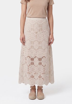Kvinde iført en beige top med korte ærmer og en lang beige blomsterblonde-nederdel med beige espadrilles med ankelrem.