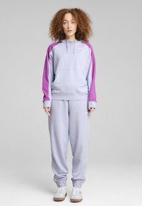 Felpa lilla con maniche viola, tasca a marsupio e cappuccio. Abbinata a pantaloni jogger lilla coordinati con strisce laterali e polsini elasticizzati.