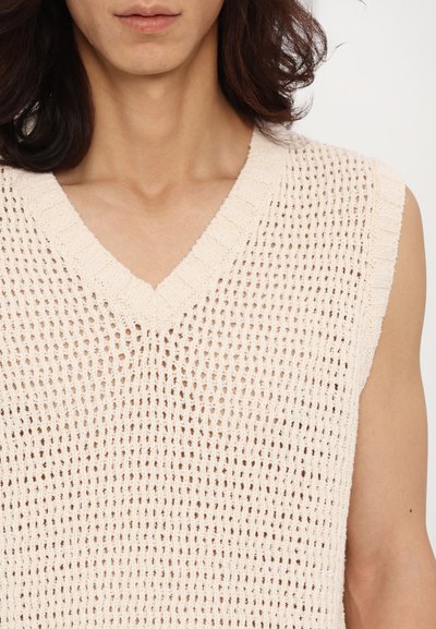Gilet tricot beige clair avec un col en V et sans manches. Le tissu présente une texture à mailles ouvertes avec un col côtelé.